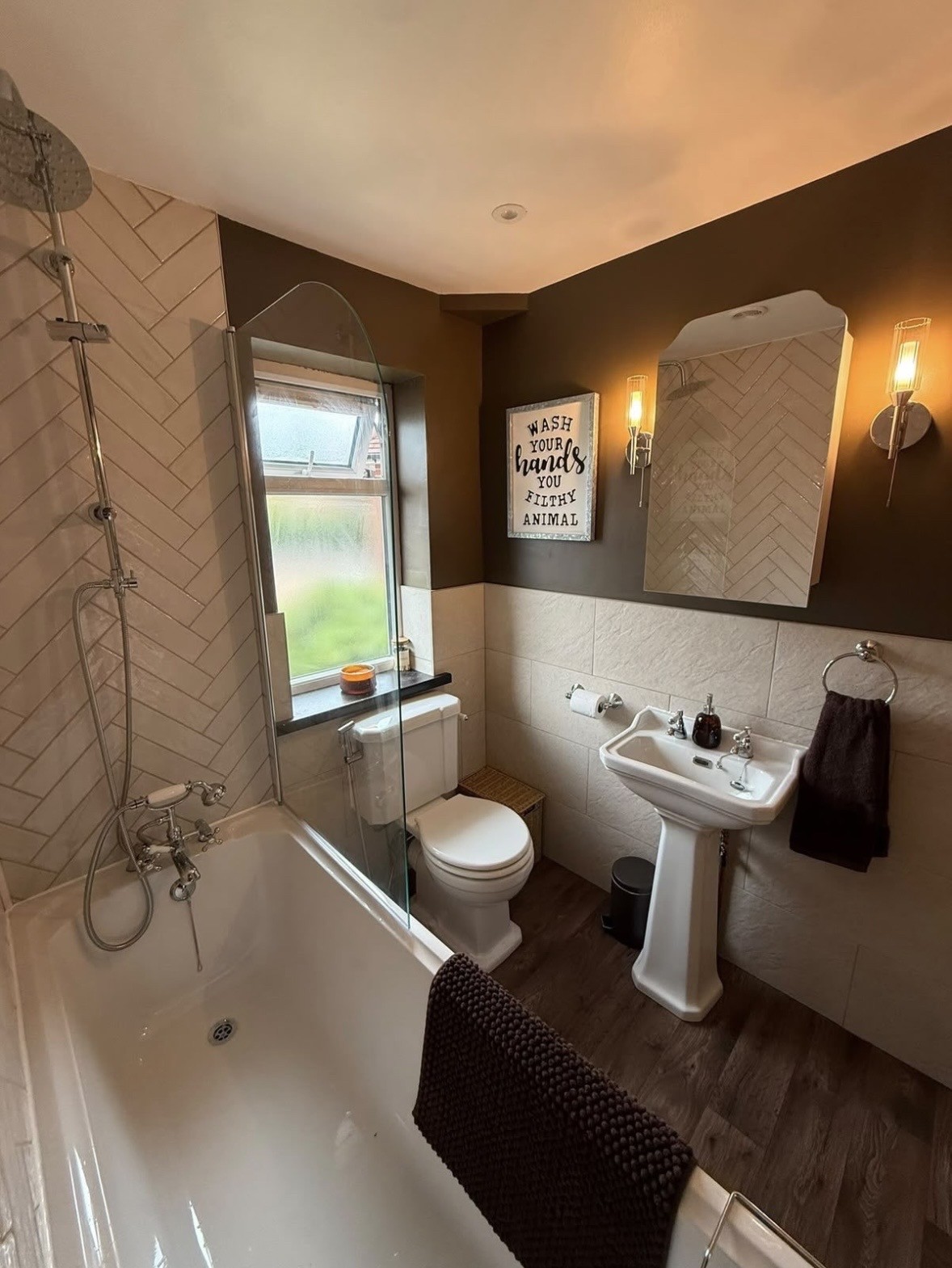 The same bathroom after the work: herringbone subway tiles, a bathtub with a chrome shower, pedestal basin and wood-effect flooring